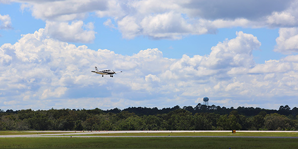 Image of an MGA plane coming down for a landing with a cloudy blue sky and lush forest in the background.